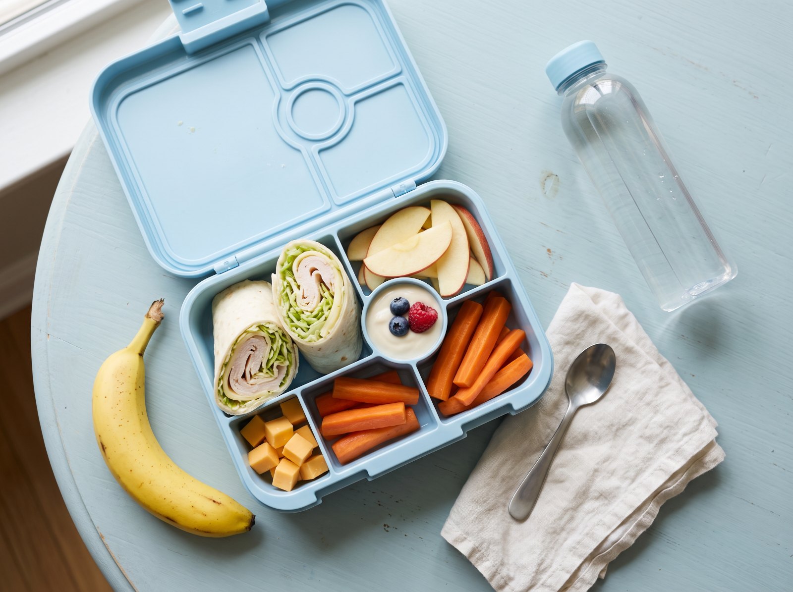An open light blue lunch box packed with a soft wrap, apple slices, yogurt with berries, carrot sticks, and cheese cubes, with a banana and water bottle, a braces-friendly school lunch.